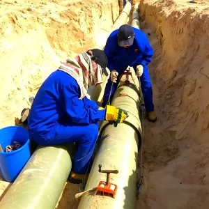 Workers installing large-diameter fiberglass round tube pipeline in desert trench, corrosion-resistant industrial fiberglass pipe for chemical/wastewater transport