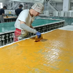 FRP grating production: Workers apply resin and glass fiber strands to a yellow mold in a factory workshop, preparing for molded fiberglass grating manufacturing.